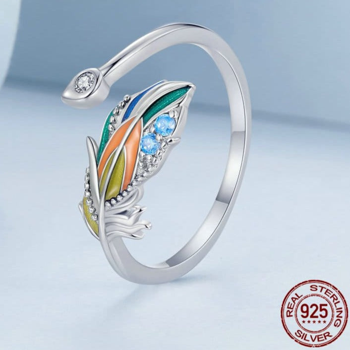 Um anel de prata 925 com uma pena colorida e pedras brilhantes na parte superior, de pé sobre um fundo azul.