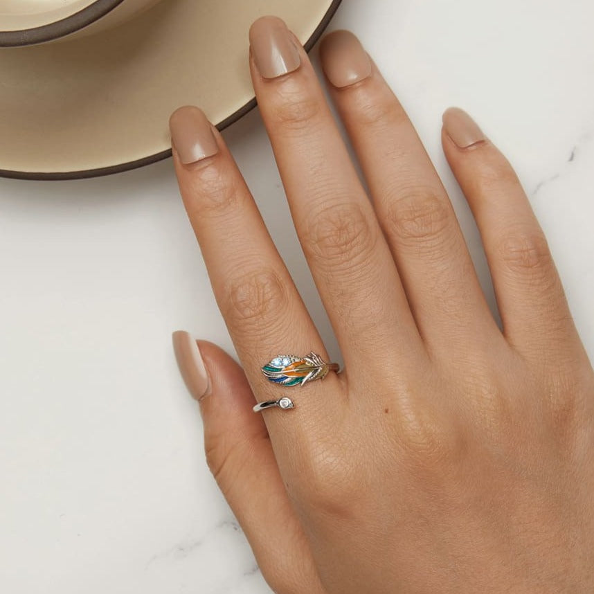 Um anel de prata 925 com uma pena colorida e pedras brilhantes na parte superior, vista de cima, no dedo de uma mão feminina aberta.