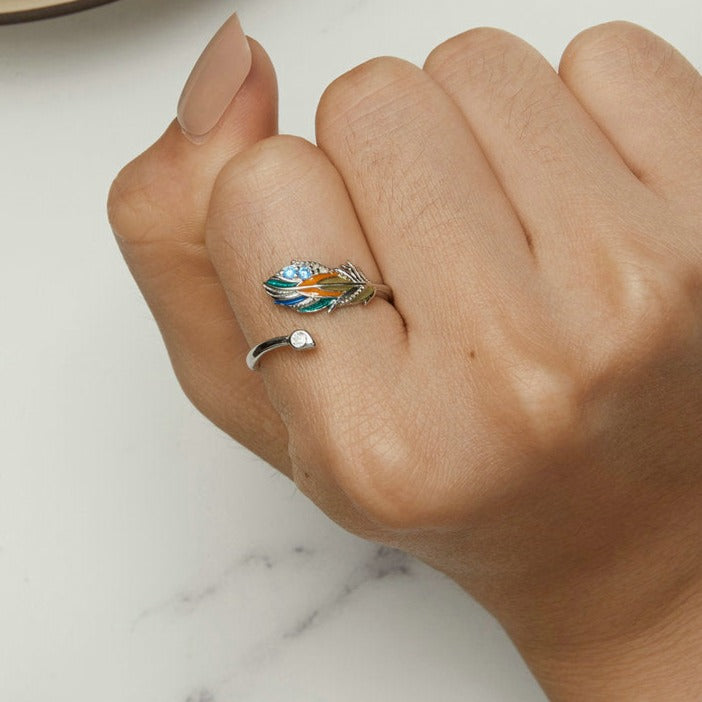 Um anel de prata 925 com uma pena colorida e pedras brilhantes na parte superior, visto de cima, no dedo de uma mão feminina.