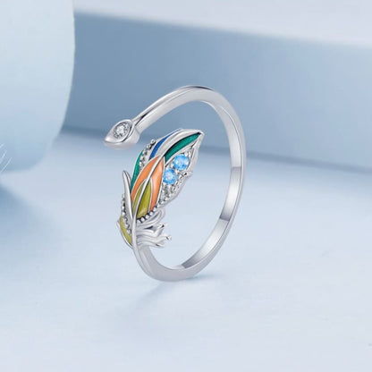 Um anel de prata 925 com uma pena colorida e pedras brilhantes na parte superior, visto de frente e de pé, sobre um funco azul.