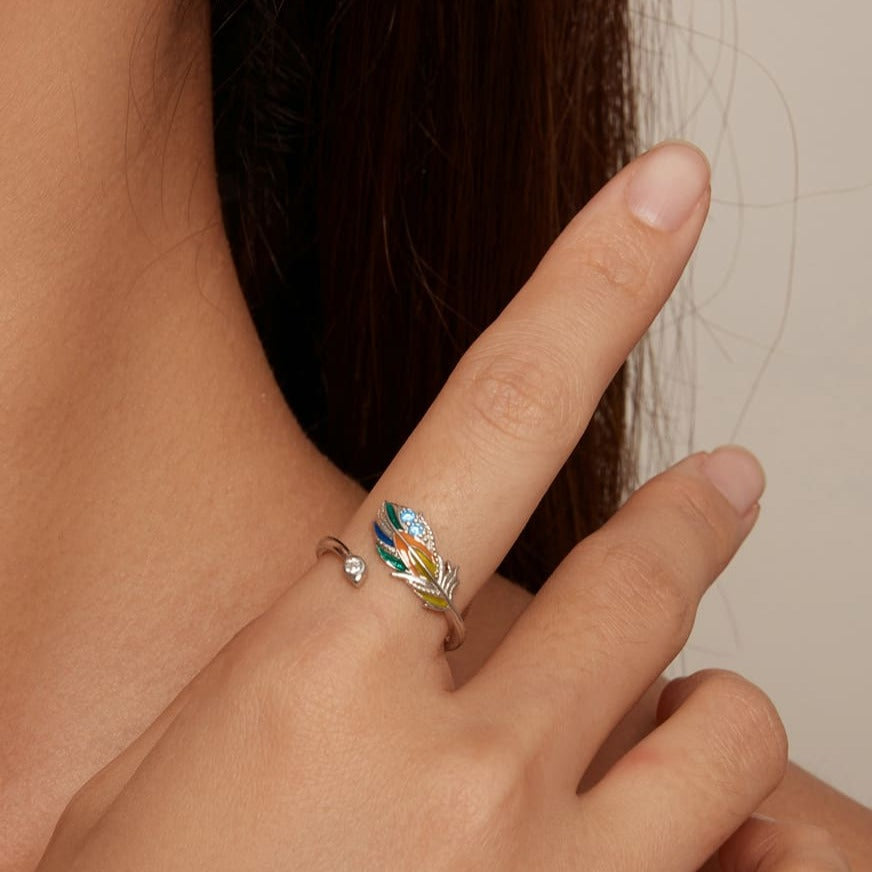 Um anel de prata 925 com uma pena colorida e pedras brilhantes na parte superior, em zoom no dedo de uma mão feminina apoiada sobre o ombro.